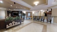 This single-floor space presents a polished and welcoming lobby lounge environment designed with both comfort and function in mind. The interior is anchored by large-format tile flooring arranged in a diagonal pattern, framed by a contrasting border that defines the central gathering area and adds a sense of refined structure to the layout.

A built-in media wall with dark wood cabinetry and a wall-mounted flat-screen television serves as a focal point on one side of the room, complemented by decorative shelving and lush greenery that adds a natural warmth to the space. Opposite, a bar-height counter with upholstered high-back stools provides a functional seating area ideal for casual meetings or social interaction.

The ceiling features a tray design with crown molding details, lending architectural depth to the room. Layered lighting — including recessed fixtures, track lighting, and flush-mount pendant lights — creates a bright and inviting atmosphere throughout. Neutral tones of cream, taupe, and warm beige are carried consistently across the walls, upholstery, and furnishings, contributing to a cohesive and sophisticated aesthetic. Additional seating with a bench and side tables offers flexible arrangements, while potted plants and decorative accents complete the well-curated ambiance of this thoughtfully designed communal space.