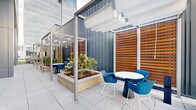 Located on Inspiration Street in Reston, VA, this thoughtfully designed outdoor terrace offers a versatile, single-level amenity space set against the backdrop of a contemporary commercial building. The terrace is organized into several distinct zones, each serving a different purpose while maintaining a cohesive modern aesthetic throughout.

One zone features vibrant green artificial turf and casual lounge seating — woven white chairs with plush cushions arranged around low side tables — creating an inviting relaxation area. A large flat-screen display mounted on a corrugated metal wall adds a media-friendly dimension to this space. An adjacent area with bistro-height tables and woven blue chairs provides options for more upright socializing or informal dining.

A covered pergola section runs along one side of the terrace, fitted with retractable sail-style shade canopies suspended from sleek steel frames. This zone incorporates raised stone planters filled with greenery, warm wood slatted privacy screens, and blue accent seating, giving it a more intimate, garden-like feel.

Broad, light-toned concrete pavers form the walkway connecting all zones, while floor-to-ceiling glazing along the building facade allows views into the interior and fills the terrace with natural light. The overall ambiance is contemporary, clean, and functional — well-suited for both casual gatherings and professional outdoor meetings.