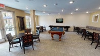 Located at 131 E Bardin Rd in Arlington, Texas, this single-floor recreational game room is thoughtfully designed to encourage social interaction and leisure. The space is anchored by a stately ornate-legged billiards table with a blue felt surface, positioned prominently at the center of the room, alongside a full-length shuffleboard table set against a patterned accent wall beneath a wall-mounted flat-screen television. Recessed lighting throughout provides consistent, even illumination across the carpeted floor, which features a neutral toned tile-pattern design. Multiple seating areas are distributed throughout the room, including dark wood tables and cushioned chairs suited for card games and board games. A dedicated chess table and a wall-mounted Scrabble board add to the variety of available activities. A built-in banquette with striped upholstery lines one wall, paired with additional dining-style tables ideal for group gatherings. Large glass doors along one side of the room flood the space with natural light and offer views of the surrounding outdoor area. Warm beige and yellow tones on the walls, complemented by tasteful framed artwork, create a welcoming and comfortable ambiance suited for community recreation and socializing.