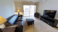 This single-floor residential unit offers a well-appointed living space designed for comfort and practicality. The living room serves as the centerpiece of the home, featuring wall-to-wall carpeting in a neutral tone that creates a warm and inviting atmosphere. Crown molding runs along the ceiling perimeter, adding a refined architectural detail to the space. Natural light fills the room generously through a large sliding glass door, which opens directly onto a private balcony — an ideal spot for outdoor relaxation with views of the surrounding neighborhood. The living area accommodates a full seating arrangement, including a leather sofa and matching accent chair, centered around a dark wood coffee table. A flat-screen television is mounted on a media console positioned opposite the seating area, creating a practical entertainment setup. Neutral-toned walls throughout contribute to a clean, open aesthetic that pairs well with the dark furnishings. Ceiling vents indicate a central climate control system for year-round comfort. The overall layout is efficient and cohesive, making good use of the available space while maintaining an uncluttered, livable environment suitable for everyday use.