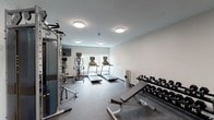 This fitness center is a thoughtfully designed, single-floor exercise space built to support a comprehensive range of workout routines. The room features durable rubber flooring throughout, providing both safety and functionality for a variety of activities. Neutral white walls and flush-mount ceiling lights create a clean, focused environment conducive to exercise.

The cardio section is well-appointed, housing multiple treadmills and an elliptical machine, each equipped with individual digital display screens. A wall-mounted flat-screen television is positioned nearby, offering entertainment during workouts. Natural light streams in through a large window along one wall, adding an airy, open quality to the space and offering a view of surrounding greenery.

For strength training, the space includes a Matrix dual cable functional trainer, ideal for a wide range of resistance exercises. A two-tier dumbbell rack stocked with a full set of free weights is positioned alongside an adjustable weight bench and an incline bench, offering versatility for both beginner and advanced users. Additional benches and adjustable seating stations are distributed throughout the room.

A mirrored wall section and a dedicated entry door with an access panel complete the space. A wall-mounted air conditioning unit ensures year-round comfort. The overall layout is open and organized, maximizing usable floor space while keeping equipment accessible and the environment uncluttered.