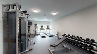 This fitness center is a thoughtfully designed, single-floor exercise space built to support a comprehensive range of workout routines. The room features durable rubber flooring throughout, providing both safety and functionality for a variety of activities. Neutral white walls and flush-mount ceiling lights create a clean, focused environment conducive to exercise.

The cardio section is well-appointed, housing multiple treadmills and an elliptical machine, each equipped with individual digital display screens. A wall-mounted flat-screen television is positioned nearby, offering entertainment during workouts. Natural light streams in through a large window along one wall, adding an airy, open quality to the space and offering a view of surrounding greenery.

For strength training, the space includes a Matrix dual cable functional trainer, ideal for a wide range of resistance exercises. A two-tier dumbbell rack stocked with a full set of free weights is positioned alongside an adjustable weight bench and an incline bench, offering versatility for both beginner and advanced users. Additional benches and adjustable seating stations are distributed throughout the room.

A mirrored wall section and a dedicated entry door with an access panel complete the space. A wall-mounted air conditioning unit ensures year-round comfort. The overall layout is open and organized, maximizing usable floor space while keeping equipment accessible and the environment uncluttered.