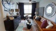 This single-floor space showcases a thoughtfully designed living area that balances bold aesthetics with functional furnishings. The room centers around a tufted cognac leather sofa paired with a dramatic tall upholstered headboard-style accent wall in grey, creating a striking focal point. A round black pedestal coffee table anchors the seating area, styled with decorative candles and metallic accents. Dark hardwood flooring is complemented by a layered vintage-style area rug in warm earth tones and turquoise, adding depth and visual interest. A dark wood dresser with silver hardware lines one wall, topped with a ceramic table lamp and decorative bowl, providing both storage and style. Natural light filters through a large window dressed with patterned dark curtains and horizontal blinds, while a ceiling fan overhead ensures comfort. A folding accent chair near the window adds a relaxed, eclectic touch. Abstract wall art contributes to the room's artistic character. The overall ambiance is sophisticated and moody, blending industrial and contemporary design elements with warm, inviting tones throughout.