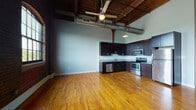 Located at 700 Stockton St in Richmond, Virginia, this single-floor loft-style unit masterfully blends historic industrial character with modern finishes. The open living and kitchen area immediately impresses with soaring ceilings framed by original heavy timber beams and rich dark wood planking overhead, complemented by exposed metal ductwork that underscores the building's industrial heritage. Warm-toned hardwood floors run throughout, reflecting natural light that filters in through large warehouse-style windows accented by original brick surrounds. The kitchen is thoughtfully appointed with dark espresso cabinetry, granite countertops, stainless steel appliances including a range, microwave, refrigerator, and dishwasher, and a double-basin sink. Industrial-style pendant and wall-mounted lighting fixtures add character throughout. A long corridor connects private rooms, each featuring the same gleaming hardwood floors and exposed wooden support columns that tie the aesthetic together cohesively. The private rooms offer neutral gray walls and ample space, while the bathroom provides clean, functional finishes including a pedestal sink, framed mirror, tiled flooring, and a full tub-shower combination. The overall ambiance is one of urban sophistication rooted in Richmond's rich architectural history.