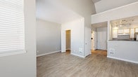 This single-floor unit is defined by its dramatic vaulted ceilings, which lend an impressive sense of height and openness throughout the living spaces. Light-toned wood-style flooring runs seamlessly across the main areas, complementing the neutral, warm-gray wall palette and crisp white trim for a clean, cohesive aesthetic.

The living area is generously proportioned, bathed in natural light through multiple large windows fitted with horizontal blinds. A standout architectural feature is the loft space accessed by a sleek black metal ladder, adding a unique vertical dimension to the open layout.

The kitchen is well-appointed with white cabinetry accented by warm wood tones, dark countertops, a stainless steel sink with dishwasher, and full appliances including a refrigerator and range. Track lighting illuminates the workspace effectively. A window above the counter brings in additional natural light.

The bedroom includes an open closet with rod storage and connects conveniently to the rest of the unit via a hallway. The bathroom features an extended vanity with dual-tone cabinetry, a dark countertop with an undermount sink, a large mirror, and wood-style flooring. The layout is efficient and well-organized throughout, making practical use of the available space while maintaining an airy, modern feel.
