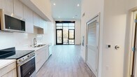 This single-floor unit showcases a contemporary design characterized by soaring ceilings, light hardwood-style flooring, and a bright, open-concept layout that seamlessly connects the kitchen and living areas. Crisp white walls and recessed lighting throughout enhance the airy atmosphere.

The kitchen is outfitted with flat-front cabinetry in a warm wood-grain finish, white quartz countertops, and a full suite of stainless steel appliances, including an electric range, built-in microwave, refrigerator, and dishwasher. A double-basin sink with a modern pull-down faucet completes the functional workspace, while upper and lower cabinetry provide ample storage.

The living area is defined by dramatic floor-to-ceiling black-framed windows and a glass door that open directly onto a private balcony, framing lush tree canopy views and flooding the interior with natural light. An additional side window further brightens the space.

The bathroom features a clean, minimalist design with light tile flooring, white walls, and accessibility-oriented grab bars, reflecting a thoughtful and inclusive approach to the overall design.