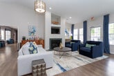 Located at 117 Teakwood Dr in Greensboro, North Carolina, this single-floor community clubhouse offers a thoughtfully designed and inviting space for residents. The entry foyer sets an immediate tone with soaring ceilings, warm hardwood-style flooring, and a striking focal wall featuring reclaimed wood panels adorned with vibrant living moss art. A rustic wood credenza with decorative blue glass accents anchors the entryway, creating a warm and welcoming first impression.

The main lounge area is generously proportioned and bathed in natural light, thanks to an abundance of large windows dressed with elegant blue drapery panels. Plush navy blue armchairs arranged around a sleek round coffee table with gold accents provide comfortable seating, while a wall-mounted fireplace with a dark stone surround adds warmth and sophistication. Built-in shelving flanks a mounted flat-screen television, offering both functional and decorative display space.

An additional sitting area features a tan leather accent chair positioned beside a bright window overlooking a pool area, complemented by a marble-top accent table and a framed full-length mirror. Throughout the space, light-toned walls, recessed lighting, and wood-look flooring create a cohesive, modern aesthetic. A service window connects to an adjacent back-of-house area, adding practical functionality to this polished community hub.