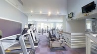 This two-floor fitness facility is designed to provide a comprehensive workout environment for users of all fitness levels. The main exercise floor is generously proportioned, offering ample space for a wide array of cardio equipment, including treadmills and stationary bikes, as well as an extensive selection of strength-training and weight machines. The layout is thoughtfully organized to ensure ease of movement between stations. Soaring ceilings lend an open, airy quality to the space, while large windows along the far wall flood the interior with natural light and provide views of the surrounding greenery. A wall of floor-to-ceiling mirrors amplifies the sense of space and allows users to monitor their form during workouts. The flooring features a durable, multi-toned carpet tile in muted purple and grey tones, providing both comfort underfoot and a polished aesthetic. Recessed lighting and track lighting ensure the space is well-lit at all times. A wall-mounted television screen is conveniently positioned for viewing during workouts. A ceiling fan adds to the comfort of the environment. The second floor is accessible via a staircase with white railing, offering additional space within the facility. Overall, the space presents a clean, bright, and professional fitness environment.