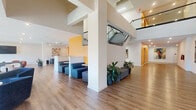 This expansive, single-floor community clubhouse offers a thoughtfully designed interior that blends modern aesthetics with functional versatility. The space is unified by warm hardwood-style flooring that runs throughout, complemented by crisp white walls and vibrant yellow accent walls that add energy and visual interest.

The grand entrance hallway is immediately striking, featuring soaring double-height ceilings, glass-railed staircases, large glass entry doors, and lush potted greenery that creates a welcoming atmosphere. Recessed lighting throughout ensures a bright, well-lit environment at all hours.

The main lounge area is generously proportioned, furnished with plush navy blue swivel chairs, channel-tufted sofas, and compact round tables ideal for both casual conversation and collaborative gatherings. A wall-mounted flat-screen TV serves as a central focal point. Floor-to-ceiling sliding glass doors open to an outdoor deck, flooding the interior with natural light and providing direct views of the surrounding community.

A separate lounge zone features rounded orange accent chairs and relaxed seating arrangements centered around another mounted display. Adjacent to this is a fully equipped kitchenette with stainless steel appliances, a large white island with bar seating, ample cabinetry, and a dedicated sink — perfect for events and everyday use. The overall ambiance is modern, vibrant, and community-oriented.