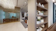 This refined single-floor space presents a seamlessly designed lobby and lounge environment that balances modern sophistication with warm, welcoming details. Upon entry, visitors are greeted by large-format light-toned tile flooring and a prominent reception desk finished in wood and gold-accented paneling, set against a dramatic textured feature wall with subtle embedded lighting. Overhead, an elaborate multi-pendant chandelier cascades across the ceiling, serving as a standout focal point throughout the space.

A semi-private meeting or conference area is enclosed by sleek glass partitions accented with vertical gold metal rods, offering visual separation while maintaining an open, connected atmosphere. Adjacent to this, a casual lounge area features plush gray seating, warm wood accent tables, a wall-mounted display screen, and a built-in media console with a beverage cooler — ideal for relaxed gatherings or informal meetings.

A dedicated display shelving unit with wood-grain finishes showcases curated decorative accessories, books, and objects, adding a boutique-like character to the space. Floor-to-ceiling sheer-draped windows flood the lounge seating area with abundant natural light and provide views of the outdoors, enhancing the open and airy ambiance. Every element of this space reflects thoughtful, high-end design with a cohesive neutral palette punctuated by gold and warm wood tones.