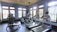 This single-floor fitness room offers a thoughtfully designed workout space finished with cool blue-grey walls, dark baseboards, and durable dark flooring suited for high-traffic exercise use. The room is lined with multiple large windows framed in warm wood trim and fitted with horizontal blinds, allowing generous natural light to fill the space while maintaining privacy. Recessed ceiling lights provide additional illumination. The cardio equipment lineup includes multiple treadmills, elliptical cross-trainers, and a stationary bike, all positioned to take advantage of the natural light and window views. A weight bench and additional strength training equipment occupy one corner, rounding out the facility's offerings. The layout is open and spacious, allowing comfortable movement between machines. The overall ambiance is clean and functional, with the light-toned walls and natural light creating an inviting, energizing atmosphere well-suited for a focused workout environment.