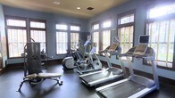 This single-floor fitness room offers a thoughtfully designed workout space finished with cool blue-grey walls, dark baseboards, and durable dark flooring suited for high-traffic exercise use. The room is lined with multiple large windows framed in warm wood trim and fitted with horizontal blinds, allowing generous natural light to fill the space while maintaining privacy. Recessed ceiling lights provide additional illumination. The cardio equipment lineup includes multiple treadmills, elliptical cross-trainers, and a stationary bike, all positioned to take advantage of the natural light and window views. A weight bench and additional strength training equipment occupy one corner, rounding out the facility's offerings. The layout is open and spacious, allowing comfortable movement between machines. The overall ambiance is clean and functional, with the light-toned walls and natural light creating an inviting, energizing atmosphere well-suited for a focused workout environment.