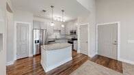 Located at Crowfield Reserve in Ladson, South Carolina, this one-bedroom, one-bathroom residence offers a well-designed single-floor layout that maximizes comfort and functionality. The open-concept kitchen serves as the heart of the home, showcasing granite countertops, a central island with pendant lighting above, crisp grey shaker-style cabinetry, and a full suite of stainless steel appliances including a refrigerator with ice maker, electric range with over-the-range microwave, and dishwasher. Rich hardwood-style flooring runs throughout the main living and kitchen areas, adding warmth and visual continuity. The spacious bedroom features plush carpeting, a ceiling fan, and a window that lets in natural light with views of the surrounding neighborhood. The bathroom is clean and well-appointed, featuring a granite-topped vanity with grey cabinetry, a tiled tub-shower combination with brushed nickel fixtures, and hardwood-style flooring that carries through from the main areas. A hallway connects the rooms efficiently, and a private entry door provides convenient exterior access. The overall aesthetic is modern and neutral, with bright white walls and consistent finishes throughout that create a cohesive, move-in-ready atmosphere.