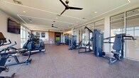 This fitness room and flex work space in Folsom, CA offers a thoughtfully designed, single-floor facility built to support a comprehensive workout experience. The generously sized exercise room is outfitted with a wide array of Matrix-brand strength machines, a full rack of dumbbells, adjustable benches, treadmills, ellipticals, stationary bikes, and a rowing machine, providing options for both cardio and strength training. Durable rubber tile flooring spans the workout area, offering both safety and functionality. The space is defined by its striking floor-to-ceiling glass wall system along one side, which floods the interior with abundant natural light and frames sweeping outdoor views of the surrounding landscape. Large ceiling fans and recessed linear LED lighting accent the crisp, modern ceiling design, contributing to a bright and comfortable atmosphere. A wall-mounted flat-screen television is positioned near the cardio zone, adding an entertainment element to workouts. Warm wood-paneled accent walls and curated artwork provide visual balance to the otherwise contemporary aesthetic. An entry hallway with glass-paneled doors connects seamlessly to the main fitness area. Adjacent to the exercise room, a spacious outdoor terrace finished with artificial turf offers an inviting extension of the facility, complete with railing-framed views of the open sky and greenery beyond.