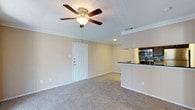 This single-floor residence offers a thoughtfully designed open-concept layout that connects the living area and kitchen in a seamless flow. The living area is finished with wall-to-wall carpet, providing a warm and comfortable underfoot feel throughout. Crown molding frames the room with a classic architectural touch, while a ceiling fan with a built-in light fixture adds both functionality and a traditional aesthetic. Recessed lighting further brightens the space, complementing the neutral, beige-toned walls that lend a clean and welcoming ambiance. The kitchen is accessible directly from the living area and features dark countertops that contrast elegantly against the lighter cabinetry and walls. Stainless steel appliances, including a double wall oven and range, are visible and underscore the kitchen's practical design. A raised breakfast bar counter serves as both a functional workspace and a subtle visual divider between the kitchen and living spaces. A paneled front door with a textured ceiling overhead completes the entryway. Overall, the space presents a cozy, functional, and well-maintained single-floor home with classic detailing throughout.