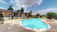 This single-story property presents a welcoming outdoor environment centered around a freeform swimming pool with clear turquoise water. The pool area is finished with a concrete surround and enclosed by a black iron fence, providing both safety and a clean aesthetic boundary. Lounge seating near the pool offers a comfortable spot to relax in the open air.

The building itself is constructed of warm brick tones with a dark brown roof, blending a classic and understated architectural style. Mature trees dot the surrounding landscape, offering natural shade and greenery that soften the overall appearance of the property. The grounds are well maintained, with planted shrubs and grass areas adding to the visual appeal.

A parking area is visible adjacent to the building, offering convenient vehicle access. The open surroundings provide an expansive feel, with wide views of the sky and nearby landscape visible beyond the fenced perimeter. The combination of the pool amenity, lush greenery, and practical layout makes this a functional and inviting property well-suited for comfortable outdoor living.