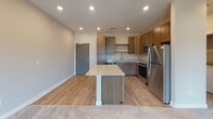 This single-floor unit showcases a thoughtfully designed open-concept layout that seamlessly connects the kitchen and living area. The kitchen is well-appointed with medium-toned wood cabinetry, light granite countertops, and a full suite of stainless steel appliances, including a refrigerator, electric range, microwave, and dishwasher. A central island with a matching granite surface provides additional prep space and serves as a natural divider between the kitchen and living area. Durable wood-look flooring runs throughout the kitchen, transitioning to plush carpet in the living space.

The living area is generously sized, featuring recessed lighting, neutral-toned walls, and a door with an adjacent window that allows natural light to fill the room and provides access to an outdoor area. The bedroom is carpeted and includes a ceiling fan with a light fixture and a window with outdoor views. The bathroom is finished with tile flooring, a granite-topped vanity with an undermount sink, and a full tub-shower combination with chrome fixtures. Clean lines and consistent finishes throughout give the unit a cohesive, contemporary feel.