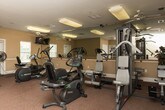 This fitness center in Decatur, Georgia offers a thoughtfully organized, single-floor layout designed to support a well-rounded workout experience. The space is carpeted in deep red tones and finished with warm beige walls, creating an inviting and energizing atmosphere throughout.

The main exercise area is generously sized, housing a variety of Precor cardio equipment including elliptical trainers and recumbent bikes, as well as a multi-station strength training machine. A wall-mounted television provides entertainment during cardio sessions, while large mirrors with a ballet barre run along one wall, visually expanding the space and supporting balance and flexibility training. Flush-mounted ceiling lights ensure the area is well illuminated throughout the day, complemented by natural light from windows along the exterior walls.

A dedicated stretching and floor exercise zone features yoga mats, a workout bench, and hand weights, providing ample space for low-impact and warm-up routines. A bulletin board with posted information and cheerful décor adds a community-oriented touch near the entrance area. A water station is conveniently accessible within the space.

The fitness center is accessible via double doors and includes clearly marked emergency exits. Overall, the space is clean, functional, and well-maintained, making it a practical amenity for residents seeking a convenient on-site exercise facility.