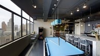 This dynamic single-floor entertainment space is designed to offer a rich social and recreational experience. At its heart sits a full-sized pool table topped with a striking blue felt surface, illuminated by a pair of distinctive pendant lights with a layered, disc-style design that serve as a bold focal point overhead. The industrial-style ceiling features exposed structural elements, track lighting, and painted ductwork, contributing to the space's modern, urban aesthetic.

Floor-to-ceiling windows line one side of the room, flooding the interior with natural light and offering sweeping views of the surrounding city skyline — a standout feature that enhances the overall ambiance. Beneath the windows, high-top tables with metal chairs provide casual seating for small groups.

An upright arcade game cabinet adds a retro entertainment element, complementing the social atmosphere. Further into the space, a dining or bar area features long communal tables with additional metal seating, suitable for group gatherings. A living wall with lush greenery adds a natural contrast to the otherwise industrial palette.

The dark tile flooring, neutral wall tones, and purposeful lighting work together to create an energetic yet polished atmosphere, making this space well-suited for recreation, socializing, and events.