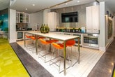 Located on Gettysburg Road in Canton, Michigan, this single-floor amenity space is designed with a contemporary aesthetic that balances functionality and comfort throughout its interconnected zones. The interior is bright and open, with recessed lighting and large windows that draw in natural light and frame views of the surrounding community and outdoor pool area.

The kitchen area is a standout feature, outfitted with sleek white cabinetry, dark countertops, a textured metallic tile backsplash, and stainless steel appliances including a wall oven and beverage refrigerators. A wall-mounted television is positioned above the backsplash, and a large white high-top table with vibrant orange stools provides a modern social dining setup.

Adjacent gathering areas feature bold, colorful furnishings, including an orange lounge chair and built-in bench seating upholstered in a multicolored wave pattern, positioned beneath windows overlooking the greenery outside. A shuffleboard table adds a recreational element to the lounge zone.

The game room area is finished with eye-catching yellow-green patterned carpet and outfitted with multiple game tables and chairs. Floor-to-ceiling glass doors open directly onto an outdoor patio, seamlessly connecting the interior to an exterior seating and entertainment area with views of the pool and surrounding buildings.