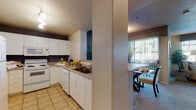 This single-floor residence offers a thoughtfully organized layout encompassing living, dining, sleeping, and utility spaces. The kitchen is laid out in a U-shaped configuration, outfitted with white cabinetry, a double sink, tile flooring, and a full suite of white appliances including an electric range, microwave, dishwasher, and refrigerator. A structural column separates the kitchen from the adjacent dining area, which features a glass-top table and vertical blinds that open toward an outdoor view.

The living room receives generous natural light through multiple windows dressed with horizontal blinds and patterned valances. Neutral wall tones and carpet flooring create a warm, relaxed atmosphere throughout the space.

Two bedrooms are present, each carpeted and featuring distinct accent walls—one bold gold and the other deep gray—providing visual contrast against otherwise neutral surroundings. Both rooms offer ample space for full bedroom furniture sets.

The bathroom includes a soaking tub and shower combination with tile surround, a white vanity with storage drawers, and a large mirror. A dedicated walk-in closet features wire shelving and carpeted flooring, providing practical and accessible storage. Overall, the layout balances functionality with a cohesive, contemporary design sensibility.