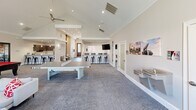 Located at 2190 Ocean Reef Place in Raleigh, NC, this single-story clubhouse facility for University Suites at Centennial offers a well-appointed amenity space designed for community engagement, productivity, and recreation. The layout encompasses a generous open-plan living area with soaring vaulted ceilings, ceiling fans, and recessed lighting throughout, creating a bright and welcoming atmosphere.

The main lounge area is anchored by a bold red billiard table and a ping-pong table, complemented by comfortable seating arrangements and a wall-mounted TV. A built-in bar-height counter with barstool seating and pendant lighting provides a casual dining and gathering spot, while a fireplace near the entrance creates a cozy focal point alongside double glass doors that invite natural light.

The dedicated office/study suite area is enclosed by sleek floor-to-ceiling glass partitions, offering multiple conference-style tables and vibrant red accent chairs — ideal for focused work or group study sessions. A separate leasing office, branded with University Suites at Centennial signage, features clean white desks and a professional layout. The facility also includes a fitness center accessible through glass doors, a kitchen area with bar seating, and a water station. Patterned carpeting and neutral tones unify the space throughout.
