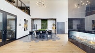 This single-floor space showcases a sophisticated contemporary design characterized by dramatic double-height ceilings that create an expansive, open atmosphere throughout. The interior seamlessly blends two distinct flooring materials — wide-plank natural hardwood and geometric dark hexagonal tile — visually defining separate functional zones within the open layout.

The main gathering area centers around a large dining table set atop the hexagonal tile field, positioned beneath a striking sculptural chandelier with an asymmetric branching design. Behind it, a framed wet bar or service area incorporates a living green wall as a dramatic focal point, flanked by pendant globe lights and recessed illumination. An adjacent floor-to-ceiling marble-clad fireplace with a see-through design adds a bold architectural statement.

The lounge area transitions naturally via the hardwood flooring, furnished with leather seating arrangements and anchored by black-framed windows that draw in generous natural light and offer exterior views. Exposed wood trusses visible in the overhead scan add structural character. Curated wall art, clean white walls, and carefully selected furnishings throughout reinforce the space's modern, upscale aesthetic.