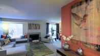 This single-story space centers around a well-appointed living area that blends contemporary design with vibrant character. A striking coral-orange accent wall serves as a focal point, adorned with large-scale artwork that adds depth and visual interest to the room. The living area features a fireplace with a clean, understated surround, anchoring the seating arrangement and creating a natural gathering point.

The space is finished with smooth, light-toned walls and neutral carpeting, providing a versatile backdrop for colorful furnishings. A bold, green patterned area rug defines the central seating zone, complementing the eclectic mix of furniture that includes a cushioned sofa, an armchair, and a sleek rectangular coffee table.

Natural light is introduced through large windows and sliding glass doors dressed in floor-length dark curtains, offering access to an outdoor area with views of greenery beyond. A decorative display ledge near the accent wall provides additional surface space, currently styled with an ornate metallic vase and floral arrangement. The overall ambiance is lively and curated, balancing bold color choices with an open, inviting layout.