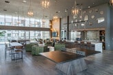 Located at 100 Zoo Ln in Santa Ana, CA, this expansive single-floor communal amenity space offers an impressive blend of modern design and upscale functionality. The standout architectural feature is its dramatic double-height ceiling, which floods the interior with natural light through floor-to-ceiling glass walls and large-pane windows that frame panoramic outdoor views of the surrounding terrace and cityscape.

Wide-plank hardwood flooring flows throughout the open layout, anchoring a series of thoughtfully arranged social zones. Elegant gold-toned chandelier pendants descend from the soaring ceiling, adding warmth and a refined aesthetic to the space. A striking feature wall clad in geometric, light-toned tile panels provides a focal point, accompanied by a mounted display screen and sculptural accent furniture.

The fully equipped kitchen bar is a central highlight, featuring an expansive granite-topped island lined with leather bar stools, stainless steel appliances, a mosaic tile backsplash, open shelving, and industrial-style pendant lighting. Decorative gold hexagonal mirrors adorn the walls above, adding an artistic dimension.

The lounge area accommodates various seating arrangements alongside recreational amenities, including a ping-pong table and game seating. Large sliding glass doors open directly onto a sunlit outdoor terrace with alfresco dining and string lighting, seamlessly connecting interior and exterior living.