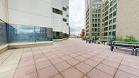 Located in the heart of Cincinnati's downtown, this rooftop terrace at 29 Seventh St W offers a thoughtfully designed outdoor space that combines open-air plazas with sheltered gathering areas. The terrace is finished throughout with large-format terracotta-toned pavers that lend warmth and visual continuity to the space.

The open-air sections feature raised planter beds lined with native grasses and greenery, bordered by wooden bench seating and sleek bollard lighting. These areas frame panoramic views of the surrounding Cincinnati skyline, with neighboring historic brick buildings and modern high-rises providing a dynamic urban backdrop.

A covered portion of the terrace provides a partially enclosed retreat, supported by concrete columns and a flat ceiling that shelters an arrangement of contemporary outdoor furniture — including lounge chairs, sofas, and low tables — creating an inviting setting for meetings or relaxation. Floor-to-ceiling glass panels along one facade reflect the surrounding cityscape, adding depth to the space.

Additional open plazas extend outward, offering generous room for gatherings or events. The terrace transitions fluidly between sun-drenched open areas and shaded covered zones, balancing functionality with an open, airy ambiance. Throughout, the architectural language is clean and modern, complementing the urban environment below.
