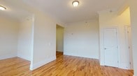 This single-floor space presents a well-maintained interior distinguished by warm honey-toned hardwood floors that extend throughout, lending a sense of continuity and natural warmth to the rooms. The walls are finished in a soft, neutral off-white tone that enhances brightness and creates an open, airy atmosphere. Crown molding lines the ceiling perimeter, adding a refined architectural detail that elevates the overall character of the space.

The main room is generously proportioned, with a flush-mount ceiling light providing central illumination. White baseboards contrast cleanly against the hardwood floors, contributing to the crisp, polished aesthetic. An open doorway leads through to an adjoining area or hallway, suggesting a practical and connected layout that supports easy movement between spaces. A closed white door on the right side of the room provides access to an additional room or storage area.

The overall ambiance is clean, light, and move-in ready, with classic architectural details that give the space a timeless quality. The neutral finishes offer a versatile backdrop suitable for a range of interior styles and furnishing choices.