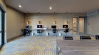 This contemporary business center is thoughtfully designed to support a range of professional needs within a single, open floor layout. The space is unified by a striking geometric carpet in grey and bold yellow, which adds visual energy and cohesion throughout.

A central conference area anchors the space, featuring a long, solid wood-toned table surrounded by ergonomic black rolling chairs — ideal for meetings, presentations, or collaborative work sessions. Nearby, a dedicated workstation area lines one wall, equipped with multiple desktop computers, task chairs, and pendant lighting fixtures suspended from the ceiling, creating a focused environment for individual work.

The walls blend smooth grey painted surfaces with a textured stone-look accent wall, adding warmth and dimension to the otherwise modern aesthetic. Curated black-and-white artwork displayed on the walls contributes a refined, gallery-like quality to the space. Recessed ceiling lights provide consistent, even illumination throughout.

Large floor-to-ceiling windows and glass doors along one side of the space flood the interior with natural light and offer views of an outdoor courtyard with mature landscaping and a patio area, creating a pleasant connection to the outdoors. A small seating lounge with upholstered chairs provides an additional casual meeting or relaxation area, rounding out this versatile and well-appointed business center.