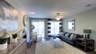 This single-floor space offers a well-appointed and thoughtfully designed interior that balances comfort with modern style. The living room serves as the centerpiece of the space, bathed in abundant natural light streaming through large windows and a sliding glass door that opens to an outdoor area, creating a seamless connection between interior and exterior. Bold navy and white striped curtains frame the windows, adding a crisp, contemporary touch to the neutral gray walls. The room features dark hardwood-style flooring that contrasts elegantly with a natural fiber area rug, anchoring the seating arrangement below. A deep navy sectional sofa accented with green and patterned throw pillows provides ample seating, complemented by a sleek white accent chair and a modern glass-top coffee table. A ceiling fan with a decorative light fixture adds both function and character overhead, while a textured ceiling lends a classic residential feel. Additional touches such as a framed wall mirror, industrial-style floor lamp, and a wood-toned dresser/console contribute to the layered, curated aesthetic. The overall ambiance is inviting and contemporary, with a neutral palette punctuated by bold accents throughout.