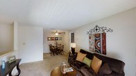 This single-floor residence offers a welcoming and functional layout designed for comfortable everyday living. The living area serves as the heart of the home, featuring plush carpeted flooring, a textured ceiling, and ample wall space ideal for artwork and décor. A comfortable seating arrangement anchors the room, complemented by warm accent pieces and decorative wall art that add character and a personal touch to the space.

The living area flows seamlessly into the dining space, creating an open-concept feel that enhances the sense of roominess. The dining area is outfitted with a ceiling fan that provides both light and air circulation, making it a practical and comfortable spot for meals. A round dining table with classic wooden chairs sits neatly within the space, reinforcing the home's warm and inviting ambiance.

Neutral wall tones and consistent carpeting throughout the shared areas create a cohesive aesthetic that is both understated and easy to personalize. The open sightlines between the living and dining zones make the space feel connected and sociable, while subtle architectural details such as clean wall transitions and recessed entryways add a sense of structure and definition to the layout overall.