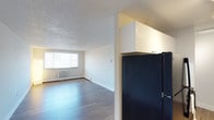 This single-floor space offers a clean, open layout designed for comfortable everyday living. The living area is bright and airy, benefiting from a large window fitted with horizontal blinds that allows natural light to fill the room. The flooring throughout is a smooth, wood-style laminate in a warm neutral tone, lending a modern and cohesive look to the interior. A baseboard heater and wall-mounted air conditioning unit line the perimeter of the living area, ensuring year-round climate control. The textured popcorn ceiling adds a classic touch overhead. Transitioning toward the kitchen area, a black refrigerator is prominently positioned beneath crisp white upper cabinetry, offering ample overhead storage. The kitchen and living areas are separated by a partial wall, creating a sense of definition while maintaining an open feel throughout the space. The overall ambiance is bright, minimalist, and well-maintained, with neutral walls and clean lines that provide a versatile backdrop suited to a variety of interior styles. This unit is ideal for those seeking a functional, straightforward living space with practical amenities and a light, welcoming atmosphere.