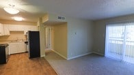 This single-floor residence features a straightforward, functional layout with a clear distinction between living and private spaces. The main living area is finished with neutral beige-toned walls and a textured popcorn ceiling, giving the space a classic, understated character. A transition from hardwood-style flooring near the kitchen area to soft carpeting in the main living zone defines the two spaces naturally without the need for barriers.

A central hallway connects the living area to the rest of the residence, offering a glimpse of additional rooms beyond. HVAC vents are integrated into the ceiling, ensuring consistent climate control throughout. Recessed architectural elements, including a boxed ceiling detail near the kitchen boundary, add a subtle layer of visual interest to the otherwise clean wall surfaces.

The kitchen area is partially visible, featuring dark appliances, including a full-size refrigerator, alongside white cabinetry. White baseboards throughout provide a crisp contrast to the warm wall tones. The overall ambiance is calm and neutral, offering a blank canvas well-suited for a variety of interior styles and furniture arrangements.