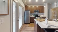 This single-floor residence showcases a cohesive mid-century modern aesthetic, blending warm walnut-toned cabinetry, light wood-style flooring, and crisp white surfaces throughout. The galley-style kitchen is a functional centerpiece, outfitted with rich dark cabinetry, white quartz countertops, a large stainless steel sink, pendant lighting, a full suite of stainless steel appliances, and a tiled backsplash. A glass-paneled entry door floods the kitchen with natural light and offers views to the outdoors. The open living area is welcoming and bright, featuring a large window that draws in ample natural light, ceiling fan, and wood-style flooring that lends warmth to the space. The two bedrooms are carpeted for comfort, each benefiting from generous windows with outdoor views and neutral, calming finishes. The bathroom is thoughtfully designed with a walnut vanity, white quartz countertop, an undermount sink, and a full tub-shower combination accented by a chevron-patterned curtain. Built-in shelving in the shower area adds practicality. Throughout the residence, consistent use of warm wood tones, modern fixtures, and clean lines creates a unified and inviting atmosphere.