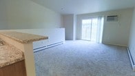 This single-floor space features an open-concept layout that seamlessly connects the kitchen and living areas. The living room is generously sized, finished with soft, light gray wall-to-wall carpeting that adds warmth and comfort underfoot. Neutral white walls keep the space bright and versatile, complementing the overall clean aesthetic.

A prominent sliding glass door dominates the far wall of the living area, allowing natural light to pour in and providing direct views of the surrounding exterior. White sheer curtains frame the door, softening the light while maintaining an airy feel.

The kitchen area is partially visible and defined by a raised countertop barrier featuring a stone-look laminate surface, creating a natural separation between the cooking and living spaces while maintaining an open feel. A white baseboard heater runs along the lower partition wall, providing efficient heating for the space.

The ceilings are standard height with a clean, simple finish, and the overall design of the space prioritizes functionality and openness. The neutral palette throughout makes it easy to personalize with furnishings and décor.