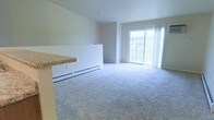 This single-floor space features an open-concept layout that seamlessly connects the kitchen and living areas. The living room is generously sized, finished with soft, light gray wall-to-wall carpeting that adds warmth and comfort underfoot. Neutral white walls keep the space bright and versatile, complementing the overall clean aesthetic.

A prominent sliding glass door dominates the far wall of the living area, allowing natural light to pour in and providing direct views of the surrounding exterior. White sheer curtains frame the door, softening the light while maintaining an airy feel.

The kitchen area is partially visible and defined by a raised countertop barrier featuring a stone-look laminate surface, creating a natural separation between the cooking and living spaces while maintaining an open feel. A white baseboard heater runs along the lower partition wall, providing efficient heating for the space.

The ceilings are standard height with a clean, simple finish, and the overall design of the space prioritizes functionality and openness. The neutral palette throughout makes it easy to personalize with furnishings and décor.