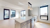 This single-floor residence showcases a sleek, contemporary design with an open-concept layout that seamlessly connects the kitchen and living areas. The kitchen is a standout feature, equipped with a large central island topped with crisp white quartz countertops, cool-toned blue-gray cabinetry with gold and brushed metal hardware, and a full suite of stainless steel appliances. A stylish pendant light fixture anchors the island, while a subway tile backsplash and additional counter space along the perimeter complete the well-appointed culinary space. Light hardwood flooring flows throughout, lending warmth and continuity across the open floor plan. The living area is bathed in natural light through expansive floor-to-ceiling windows framed in dark trim, offering sweeping views of the surrounding urban skyline. Built-in shelving adds a functional architectural touch to the space. The primary bathroom features a glass-enclosed walk-in shower, a dual-sink vanity with white countertops, matte black fixtures, and direct access to a walk-in closet. A second full bathroom offers a single-sink vanity with matching blue-gray cabinetry, matte black hardware, and warm hardwood flooring. The overall ambiance is clean, bright, and modern throughout.