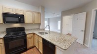This single-floor, 2-bedroom, 2-bathroom residence offers a well-designed open-concept layout that seamlessly connects the kitchen and living areas. The kitchen is a functional centerpiece, featuring light maple cabinetry, granite countertops with subtle veining, and a peninsula with an undermount sink that opens directly to the living space — ideal for both everyday living and entertaining. Black appliances, including an electric range, built-in microwave, and dishwasher, provide a clean contrast to the warm tones of the cabinetry and counters. Hardwood-style flooring runs through the kitchen, adding warmth underfoot. Track lighting overhead ensures the space is well-lit throughout. The adjacent living area is bright and airy, benefiting from multiple windows that invite natural light. A walk-in closet is visible off one of the bedrooms, hinting at ample storage throughout the home. Neutral wall tones create a versatile backdrop throughout, allowing the architectural details and finishes to take center stage. The overall ambiance is clean, modern, and move-in ready.