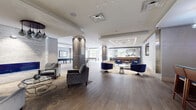 This single-floor amenity space is designed with a contemporary aesthetic, combining open-plan living areas with distinct functional zones. Wide-plank hardwood flooring runs throughout, unifying the space under recessed and linear LED lighting that highlights architectural details at every turn.

The expansive main lounge is anchored by a striking linear gas fireplace set against a textured stone accent wall, with curated seating arrangements in grey and navy that invite relaxed conversation. Structural columns clad in dark grey paneling divide the space gracefully without interrupting the open flow.

A dedicated billiards area sits adjacent to the lounge, bathed in natural light from large perimeter windows that offer views of the surrounding outdoor greenery. Nearby, a full-length bar with a white quartz countertop and a row of upholstered barstools serves as an elegant social hub, complete with a wall-mounted display and direct access to the outdoors.

A private media lounge offers an intimate retreat, featuring tufted leather seating, a wall-mounted television on a textured panel wall, and a crystal chandelier overhead. The lobby entry area provides a refined welcome with plush grey armchairs, curated artwork, and a modern pendant light fixture positioned near the elevator bank.
