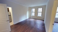This single-floor residence offers a clean, modern aesthetic anchored by rich, dark hardwood flooring that runs consistently throughout the main living areas and bedrooms. Crown molding adds a refined architectural detail along the ceilings, complementing the crisp white trim and warm neutral walls throughout.

The main living area is generously proportioned, featuring dual windows that frame views of the surrounding urban streetscape, allowing natural light to fill the space. Baseboard heating units line the walls beneath the windows, ensuring year-round comfort. The open layout connects seamlessly to additional rooms via wide doorways, enhancing the overall sense of flow.

The kitchen is efficiently designed and fully equipped with stainless steel appliances, including a full-size refrigerator, an electric range with oven, and an over-the-range microwave. Dark espresso cabinetry provides ample storage above, while a mosaic tile backsplash adds a stylish accent. Light-toned flooring in the kitchen contrasts pleasantly with the darker finishes elsewhere.

The bedroom is a comfortable, well-lit room with its own window offering city views, along with a closet and the same warm hardwood flooring found throughout. The bathroom is finished with classic white square tile work surrounding the tub and shower area, chrome fixtures, a vanity with dark cabinetry, and clean white countertops, creating a simple and functional space.