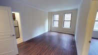 This single-floor residence offers a clean, modern aesthetic anchored by rich, dark hardwood flooring that runs consistently throughout the main living areas and bedrooms. Crown molding adds a refined architectural detail along the ceilings, complementing the crisp white trim and warm neutral walls throughout.

The main living area is generously proportioned, featuring dual windows that frame views of the surrounding urban streetscape, allowing natural light to fill the space. Baseboard heating units line the walls beneath the windows, ensuring year-round comfort. The open layout connects seamlessly to additional rooms via wide doorways, enhancing the overall sense of flow.

The kitchen is efficiently designed and fully equipped with stainless steel appliances, including a full-size refrigerator, an electric range with oven, and an over-the-range microwave. Dark espresso cabinetry provides ample storage above, while a mosaic tile backsplash adds a stylish accent. Light-toned flooring in the kitchen contrasts pleasantly with the darker finishes elsewhere.

The bedroom is a comfortable, well-lit room with its own window offering city views, along with a closet and the same warm hardwood flooring found throughout. The bathroom is finished with classic white square tile work surrounding the tub and shower area, chrome fixtures, a vanity with dark cabinetry, and clean white countertops, creating a simple and functional space.