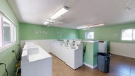 This shared laundry facility is designed with both functionality and comfort in mind. The single-floor space features a clean, inviting interior with light green walls accented by white wainscoting, creating a bright and welcoming atmosphere. Decorative lettering spelling out "Wash, Dry & Fold" adds a playful touch to the walls, reinforcing the room's dedicated purpose.

The facility is equipped with a row of top-loading washing machines and multiple dryers, providing ample capacity for simultaneous use. Spacious white folding counters line the walls, offering convenient surfaces for sorting and folding laundry. A window allows for natural light to supplement the overhead fluorescent lighting, which ensures the space remains well-lit at all hours.

Ceiling fans are mounted throughout the room, promoting air circulation and maintaining a comfortable environment during use. The tile flooring is easy to clean and maintain, contributing to the overall practicality of the space. A waste bin is conveniently positioned near the machines for easy disposal. The layout is thoughtfully organized to allow users to move freely between machines and folding areas, making the laundry process as efficient and straightforward as possible.