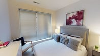 The bedroom is thoughtfully designed with a clean, contemporary aesthetic that balances comfort and style. The room is anchored by a generously sized bed with a tufted, upholstered grey headboard accented with nailhead trim detailing, dressed in crisp white linens and layered with an assortment of decorative pillows in neutral and patterned fabrics. A large window fitted with wide horizontal blinds occupies a prominent wall, allowing natural light to filter softly into the space while maintaining privacy. The neutral, warm-toned walls provide a calm backdrop, complemented by a bold abstract artwork in deep crimson and grey tones mounted above the headboard, adding a striking visual focal point. A compact nightstand sits beside the bed, topped with a simple white table lamp that contributes to the room's warm, ambient lighting. A wooden-framed accent chair with light upholstery occupies one corner, offering additional seating. The carpeted flooring adds warmth and texture underfoot, enhancing the overall sense of comfort. The space is efficiently laid out, making excellent use of its footprint while maintaining an uncluttered, inviting atmosphere.