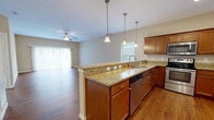 This single-story home offers a functional and comfortable layout across two bedrooms and two bathrooms. The open-concept kitchen and living area forms the heart of the home, where rich hardwood-style flooring, warm neutral tones, and ample natural light from large windows create a welcoming atmosphere. The kitchen is well-equipped with warm-toned wood cabinetry, granite countertops, a peninsula with pendant lighting and an integrated sink, and a full suite of stainless steel appliances. The adjacent living room features a ceiling fan and sliding glass access, adding to the bright, airy feel of the shared space. The generously sized primary bedroom includes a walk-in closet fitted with wire shelving and carpeted flooring. Both bathrooms echo the home's design language with granite-topped vanities and wood cabinetry; the primary bath includes a glass-enclosed walk-in shower and hardwood-style flooring. A dedicated laundry room provides a utility sink, washer and dryer hookups, and overhead wire shelving for additional storage. The attached garage offers substantial vehicle and storage space, complete with a paneled door featuring transom windows. Outside, the home presents a classic vinyl-sided exterior with a well-maintained lawn and a white privacy fence.