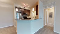 This single-floor unit features an open-concept design that seamlessly connects the kitchen and living areas, creating a spacious and functional living environment. The kitchen is a standout feature, showcasing rich dark-wood cabinetry that provides ample storage, paired with granite countertops that extend across a curved breakfast bar — ideal for casual dining or entertaining. Stainless steel appliances, including a refrigerator, microwave, and range, complement the modern aesthetic.

Track lighting above the kitchen and pendant lights above the breakfast bar provide layered illumination, enhancing both the functionality and ambiance of the space. Light wood-tone flooring runs throughout, adding warmth and continuity between rooms. The neutral wall tones keep the space feeling bright and open.

A hallway visible from the kitchen leads to the bedroom and bathroom, offering a logical and private layout. The unit's design balances practicality with contemporary style, making it a comfortable and well-equipped living space suited for individuals or couples seeking modern, move-in-ready accommodations.