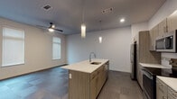 Located along W Pauline Whitaker Pkwy in Rogers, Arkansas, this single-floor unit delivers a clean, modern aesthetic with thoughtfully designed living spaces. The open-concept kitchen serves as the central hub of the unit, anchored by a sizable center island with a white quartz countertop, integrated sink, and warm wood-tone cabinetry. Pendant lighting descends from above, complementing recessed fixtures throughout. Stainless steel appliances — including a refrigerator, range, and microwave — complete the contemporary kitchen setup. The flooring in the main living and kitchen areas features a patterned tile in muted tones, while bedrooms are finished with plush, neutral-toned carpet. Each bedroom benefits from natural light via tall, blinds-fitted windows and ceiling fans for climate comfort. The bathroom is appointed with a tiled tub-and-shower surround, a wood-tone vanity with a light countertop, large mirror, and open dark-wood shelving for storage. A dedicated laundry area accommodates a stacked washer and dryer unit alongside built-in closet rod storage. The overall palette — whites, grays, and warm wood tones — creates a cohesive, contemporary atmosphere throughout.