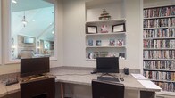 Located at 4311 Camden Cir in Dublin, Ohio, this single-floor home offers a thoughtfully designed interior with functional and stylish spaces throughout. A standout feature is the dedicated home office, which is equipped with a generous built-in desk topped with granite-style countertop surface, providing ample workspace for productivity. Integrated wall shelving above the desk offers organized display and storage space, currently home to framed awards and media collections. A window adjacent to the workspace offers a direct sightline into a naturally lit living area, which features vaulted ceilings, warm pendant lighting, and light-toned furnishings that contribute to an open, welcoming ambiance. The living space benefits from abundant natural light, with tall windows that draw in daylight and enhance the airy feel of the room. The home's interior palette is neutral and clean, with white walls and trim that create a bright, cohesive aesthetic throughout. The overall layout reflects a single-story design that promotes easy flow between living and working areas, making it well-suited for both everyday living and functional home use.